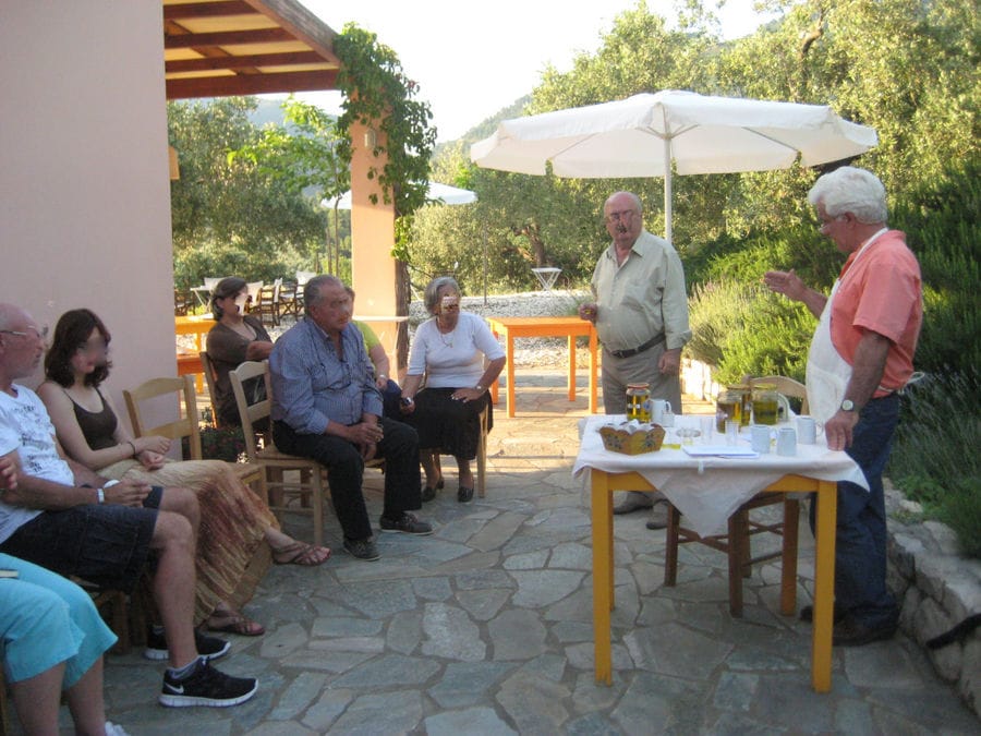 a group of tourists listening to a man giving a olive tasting at 'Eleonas' outside